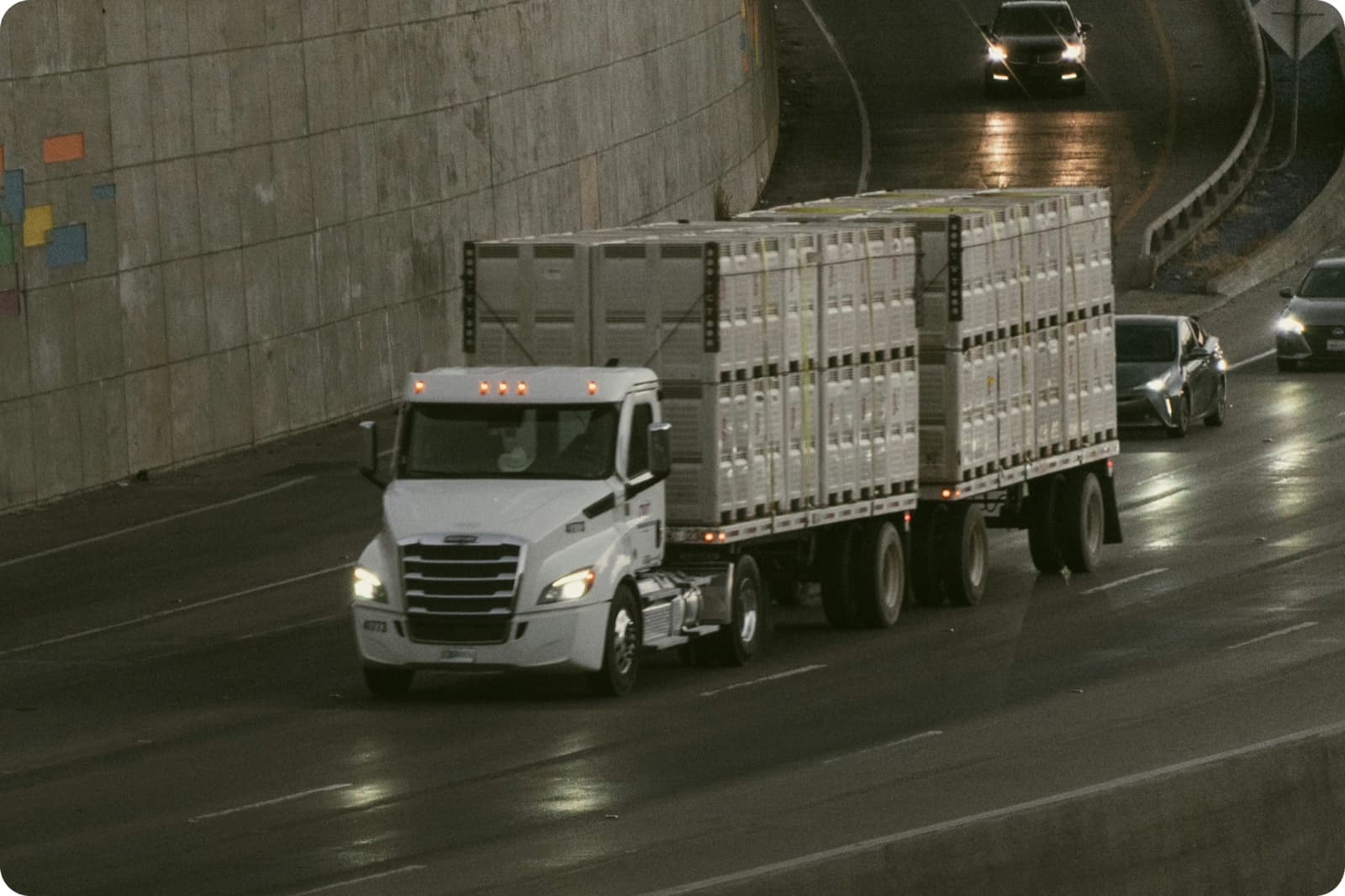 A white semi-truck hauling two connected trailers stacked with crated freight, driving through an underpass on a wet highway at dusk