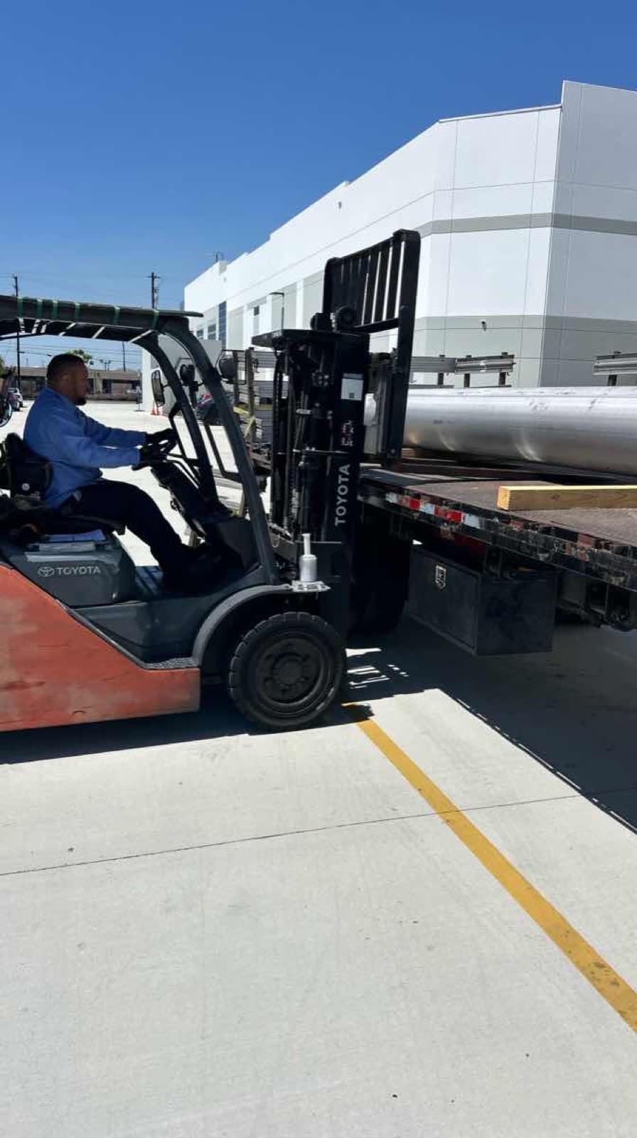 Orange Toyota forklift loading bundles of aluminum cylindrical tubing onto a Curri flatbed truck in a sunny industrial yard, ready for a same-day Moffett-equipped jobsite delivery