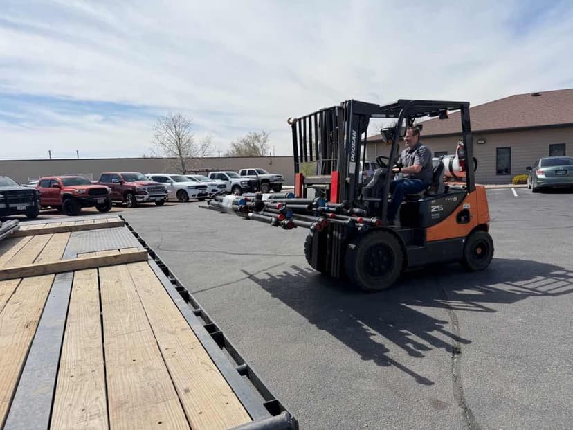 Orange Doosan forklift carrying long pipes near a Curri flatbed for an industrial-supply jobsite drop-off