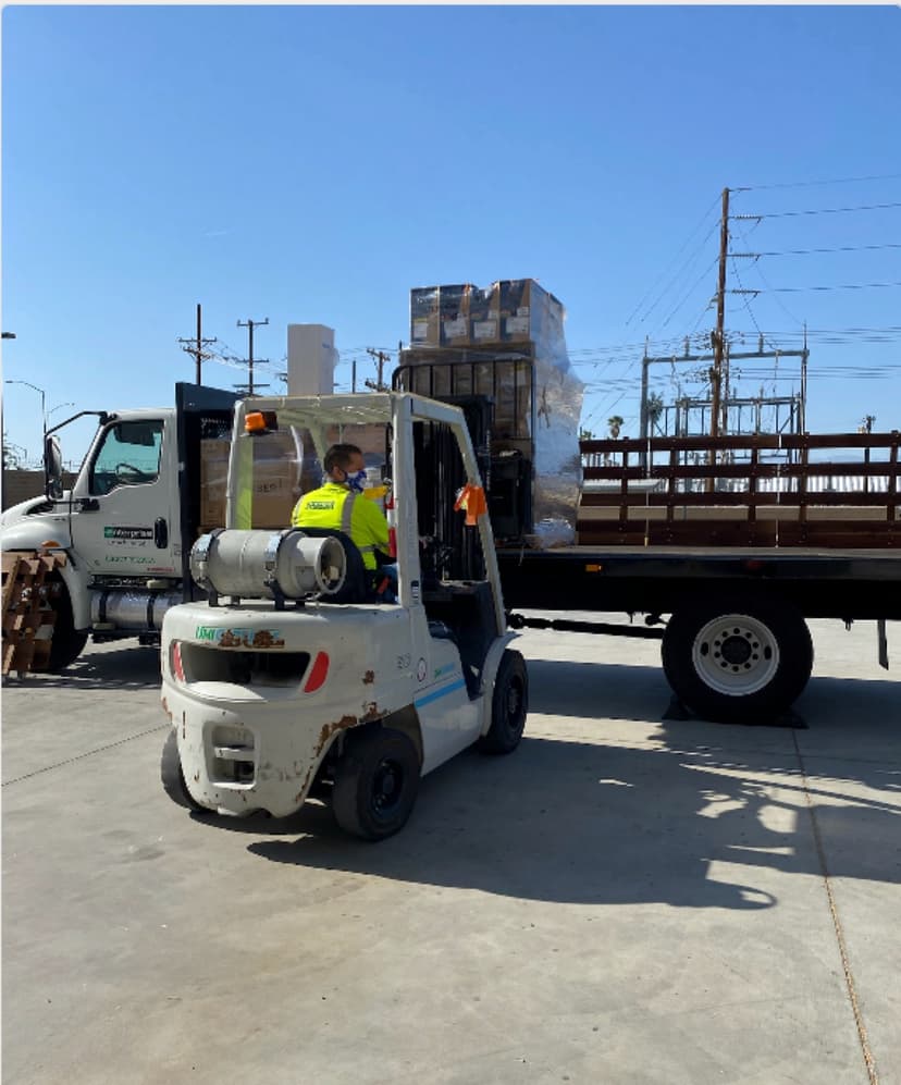 White forklift loading pallets onto a stake-sided flatbed truck for a Curri Moffett-style jobsite delivery