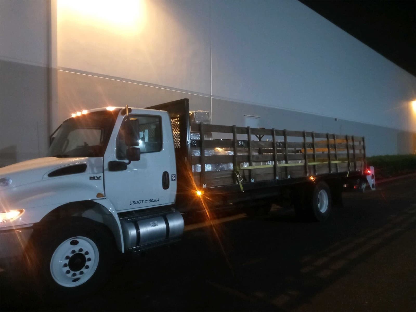 Night shot of a white Curri stakebed alongside a warehouse with strapped lumber bundles ready for an early morning route