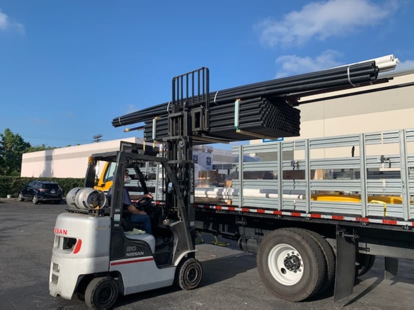 Forklift loading a black pipe bundle onto a galvanized stakebed for a Curri plumbing-supply distribution run