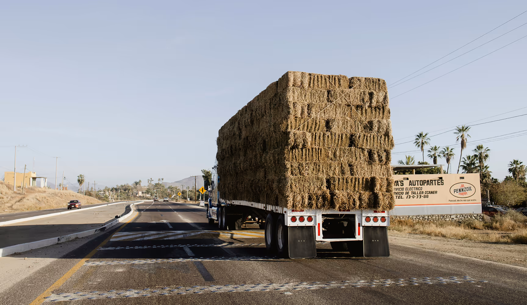 Flatbed semi hauls a towering load of hay bales down a rural highway, representing the agricultural freight and ag-supply deliveries Curri coordinates between co-ops, dealers, and farms.