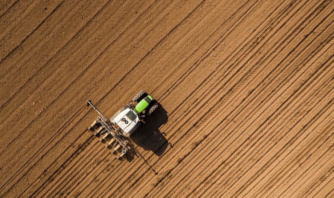 Aerial view of a green tractor pulling a seeder across a freshly tilled brown field, the kind of farm operation Curri supports with same-day delivery of feed, seed, fertilizer, and ag-supply parts.