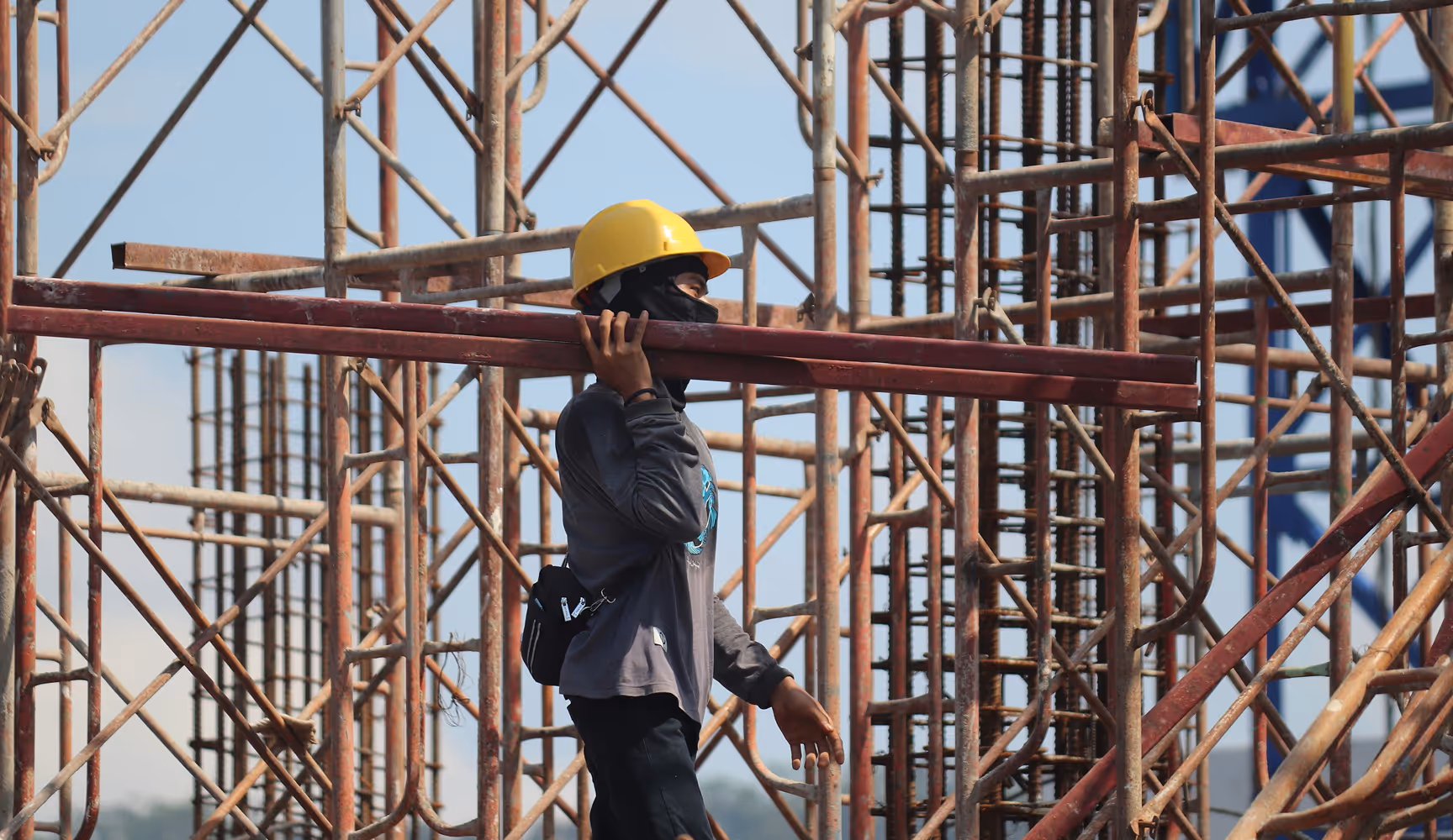 Construction worker in a yellow hard hat shoulders a steel beam through a maze of scaffolding, representing the drywall, rebar, fasteners, and lumber Curri moves to contractors and jobsites on demand.
