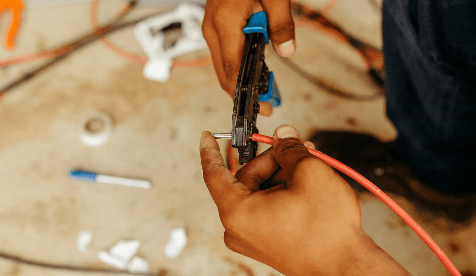 Electrician's hands strip insulation off a red electrical cable with wire strippers on a jobsite floor—the kind of wire, conduit, and gear Curri delivers from electrical distributors to contractors same-day.