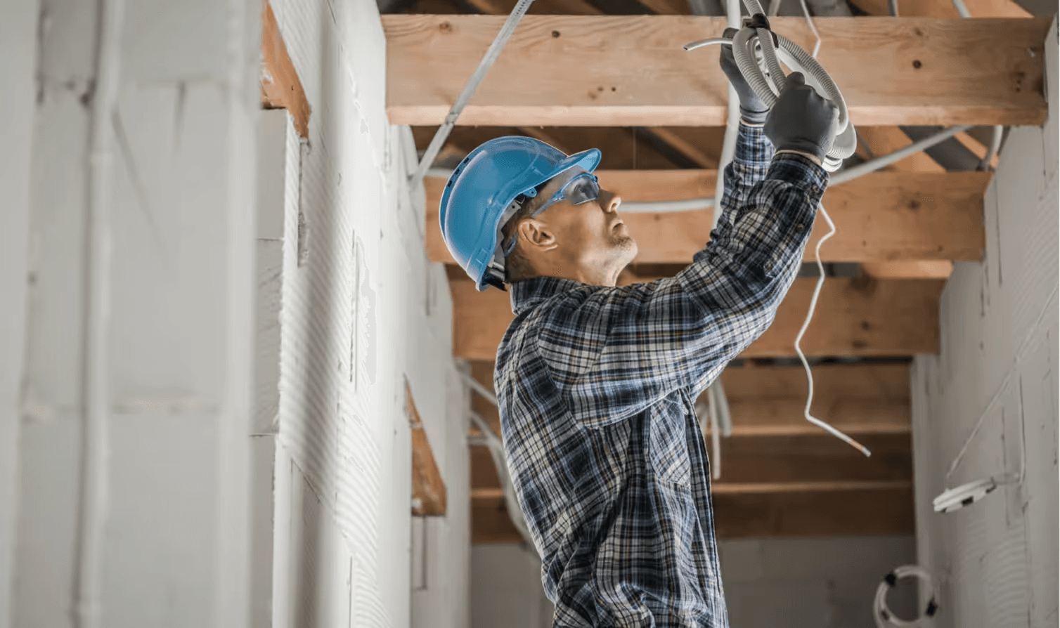 Licensed electrician in a blue hard hat runs wire through ceiling framing in an unfinished room, the kind of jobsite where Curri delivers conduit, wire spools, switchgear, and breaker panels same-day.
