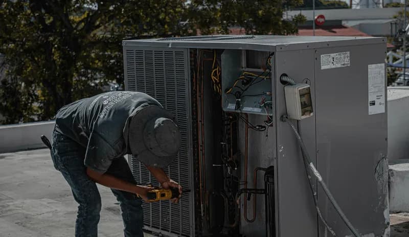 HVAC technician leans over a rooftop condenser unit to service the compressor and refrigerant lines, representing the heat pumps, condensers, and parts Curri delivers to techs and contractors same-day.