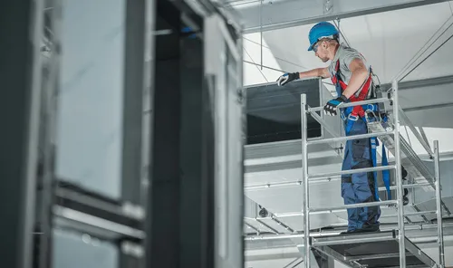 HVAC technician on rolling scaffolding installs a long run of sheet-metal ductwork overhead in a mechanical room, the kind of install Curri supports with same-day delivery of ducts, fittings, and supplies.