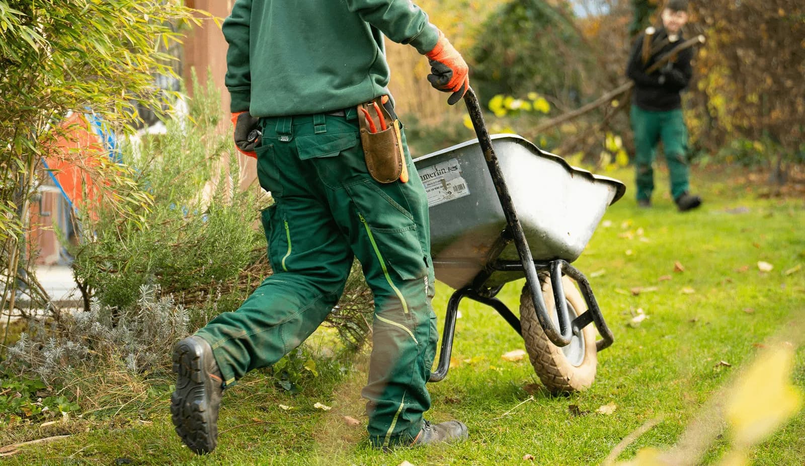 Landscaper in green work gear pushes a wheelbarrow across a lawn with a tool apron at his waist, representing the mulch, sod, pavers, and hardscape Curri delivers to crews and supply yards same-day.
