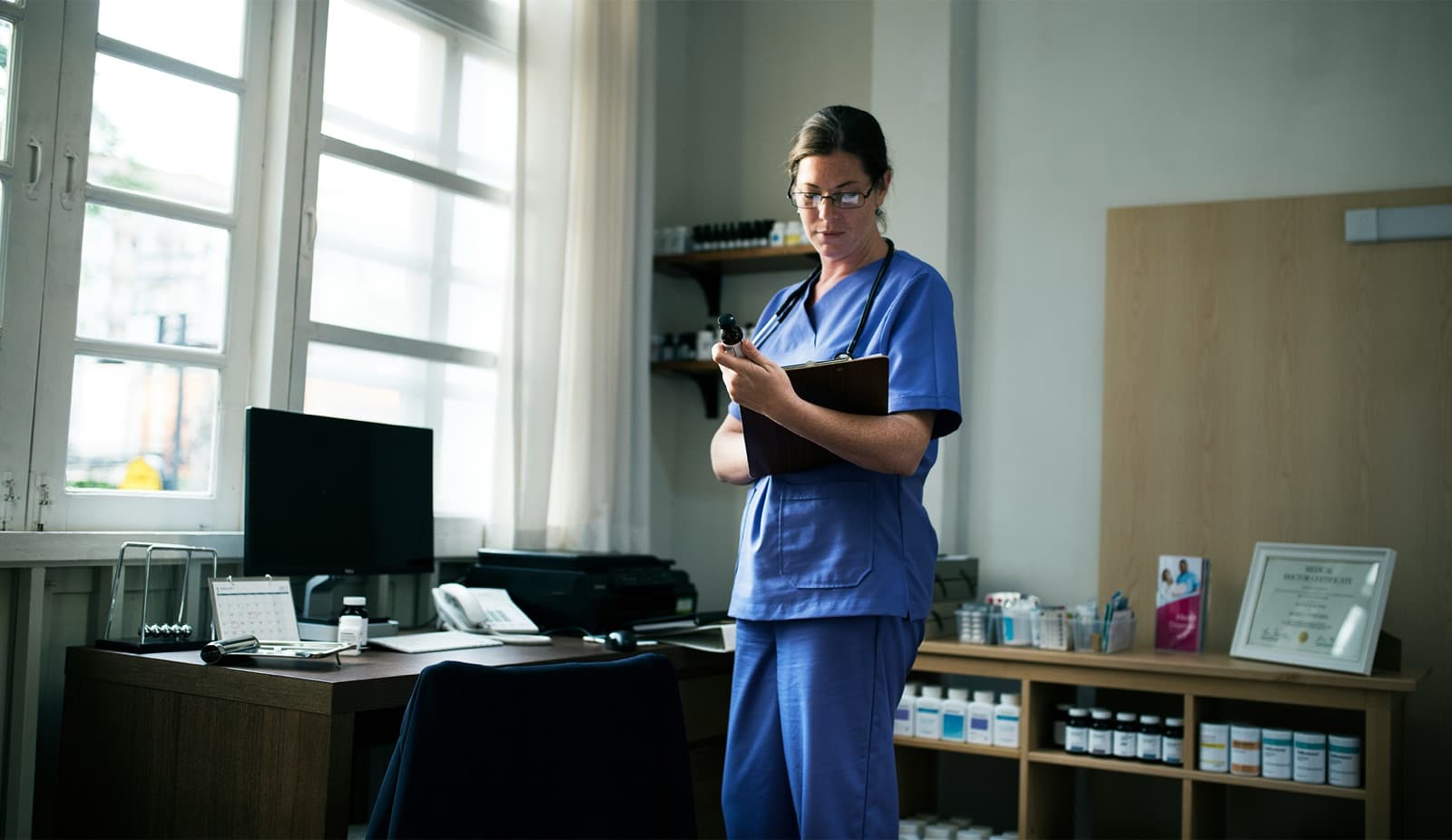 Medical professional in blue scrubs stands in a clinic office holding a clipboard next to shelves of labeled bottles, representing the PPE, pharmaceuticals, and clinical supplies Curri delivers with compliance in mind.