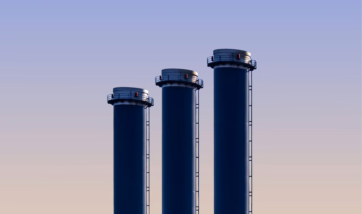 Three tall navy industrial smokestacks rise against a gradient dawn sky, representing the upstream, midstream, and well-site operations Curri supplies same-day with pipe, fittings, and MRO parts.
