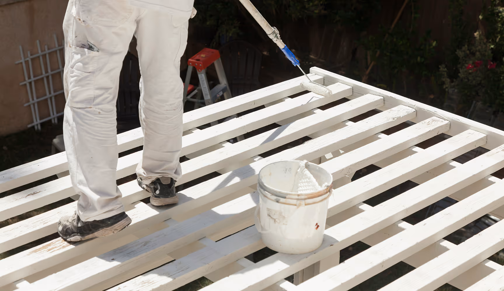 Painter in white coveralls rolls fresh white paint across a wooden pergola frame with a bucket at his feet, representing the architectural and commercial coatings Curri moves from suppliers to pro painters.