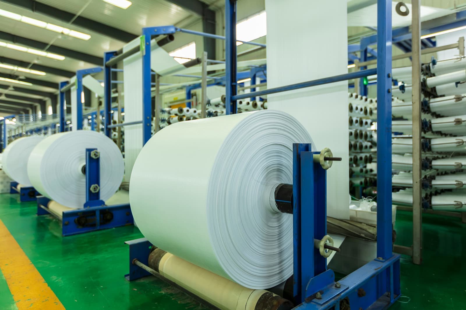 Rows of industrial looms and spindles produce woven polymer fabric on massive white rolls inside a plastics factory, the kind of extrusion and resin output Curri moves to converters and injection molders.
