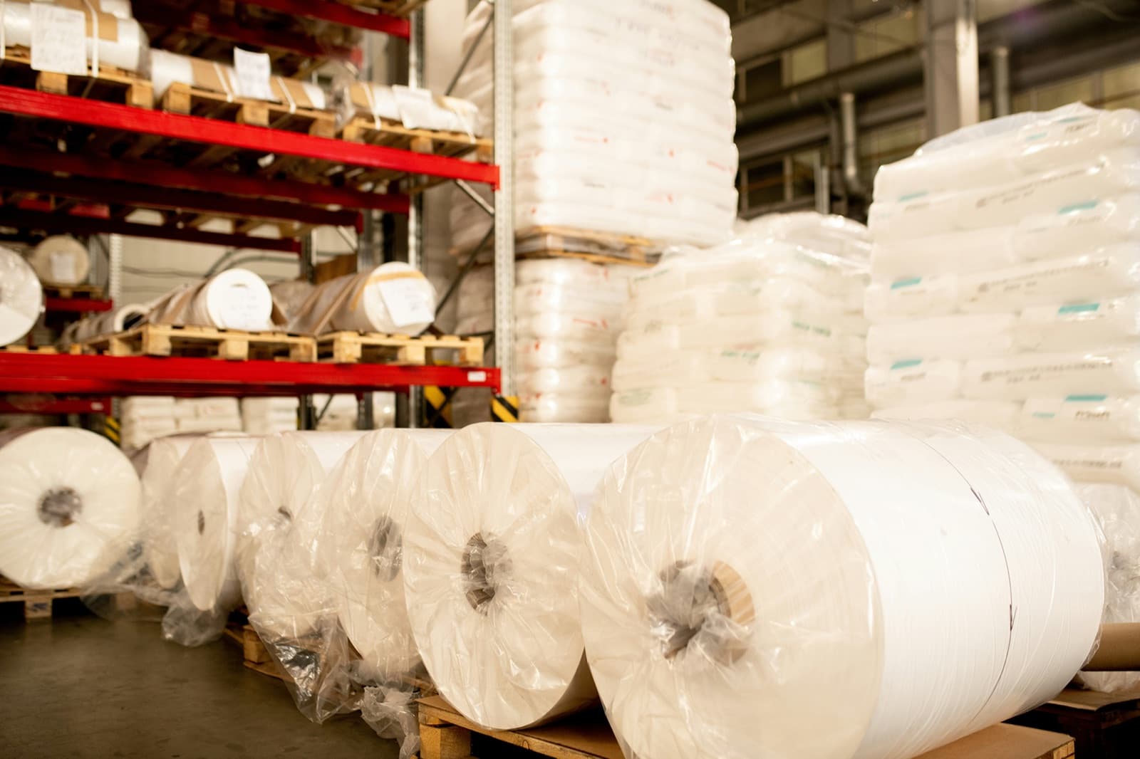 Plastics warehouse stacked with giant rolls of polymer film, pallets of resin in super sacks, and racks of raw material, representing the resin, pellets, and film Curri delivers same-day nationwide.