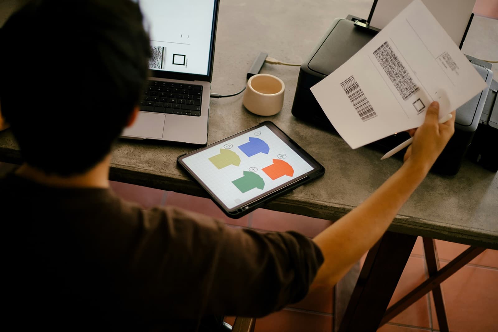 Designer reviews a tablet showing colorful house-shaped icons beside a laptop and freshly printed QR-coded sheets, representing the DTG, DTF, apparel, and signage orders Curri delivers nationwide.