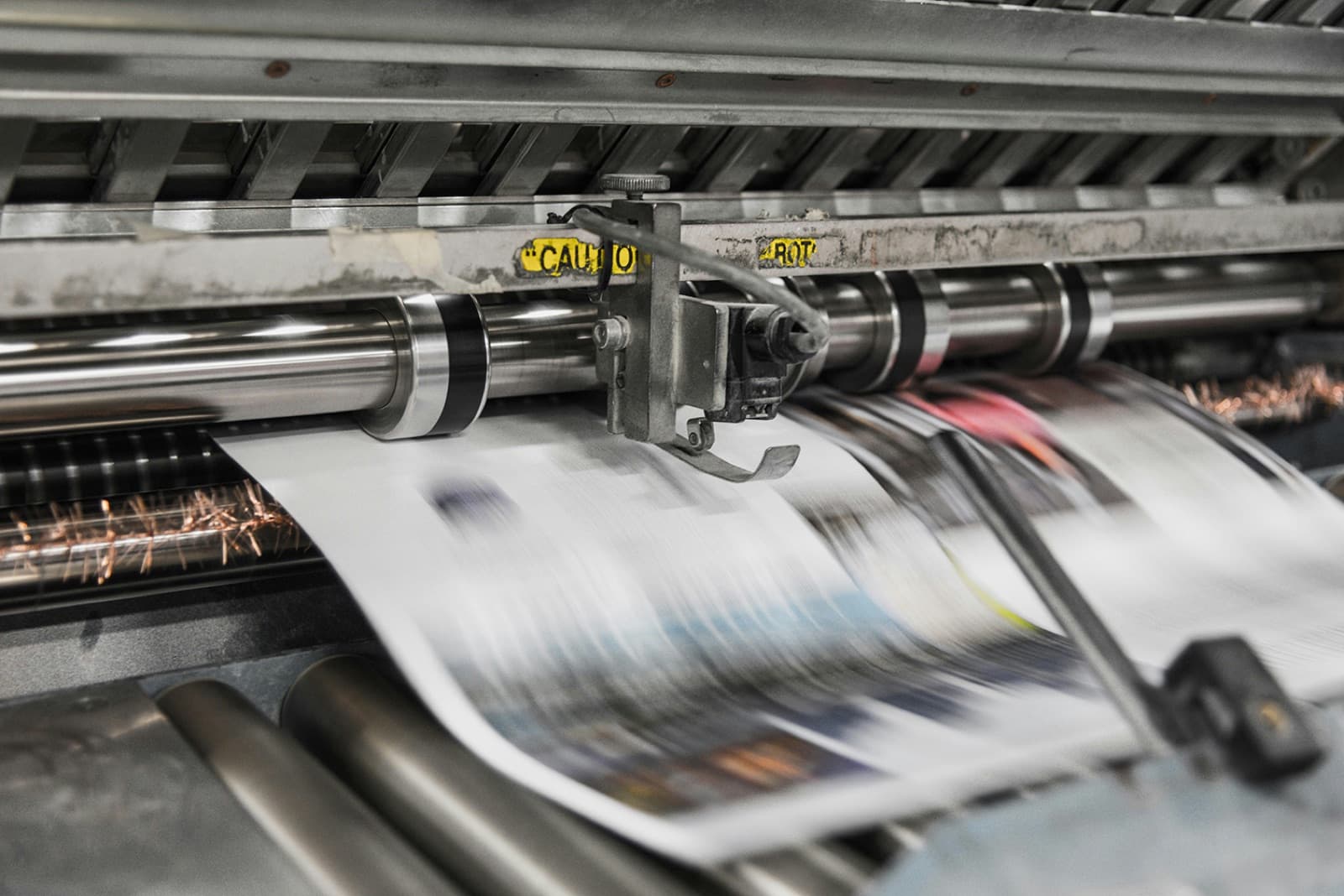 Close-up of an offset printing press running full-color pages through inked rollers with a blur of sparks, the kind of POD production Curri supports with same-day apparel, merch, and signage delivery.