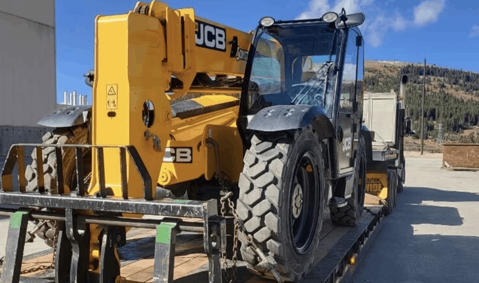 Yellow JCB telehandler loaded and chained to a flatbed hauler in a rental yard, the kind of aerial lift and heavy machine Curri coordinates rental delivery and pickup for fleets nationwide.
