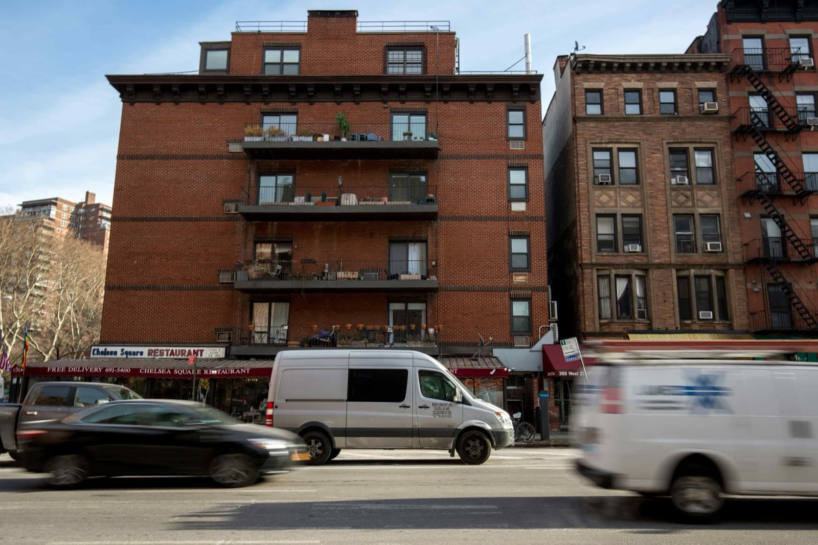 Cargo vans and cars stream past a brick New York apartment building with a corner restaurant, the dense urban last-mile environment where Curri moves BOPIS and buy-online-deliver-from-store orders for retailers.