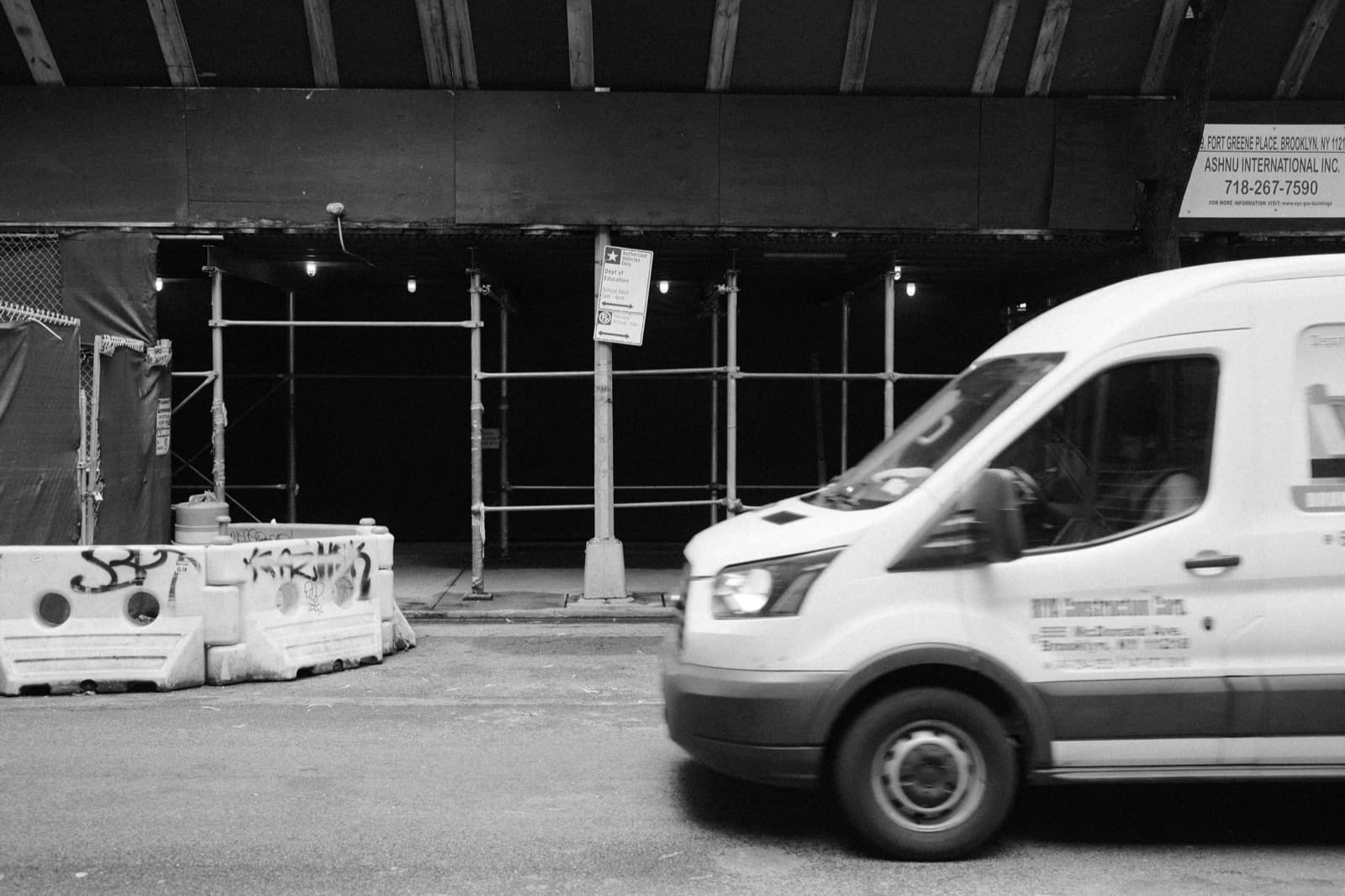 Black-and-white street shot of a white cargo van passing a scaffolded storefront in Brooklyn, the kind of urban last-mile delivery Curri powers for nationwide retailers fulfilling same-day orders.