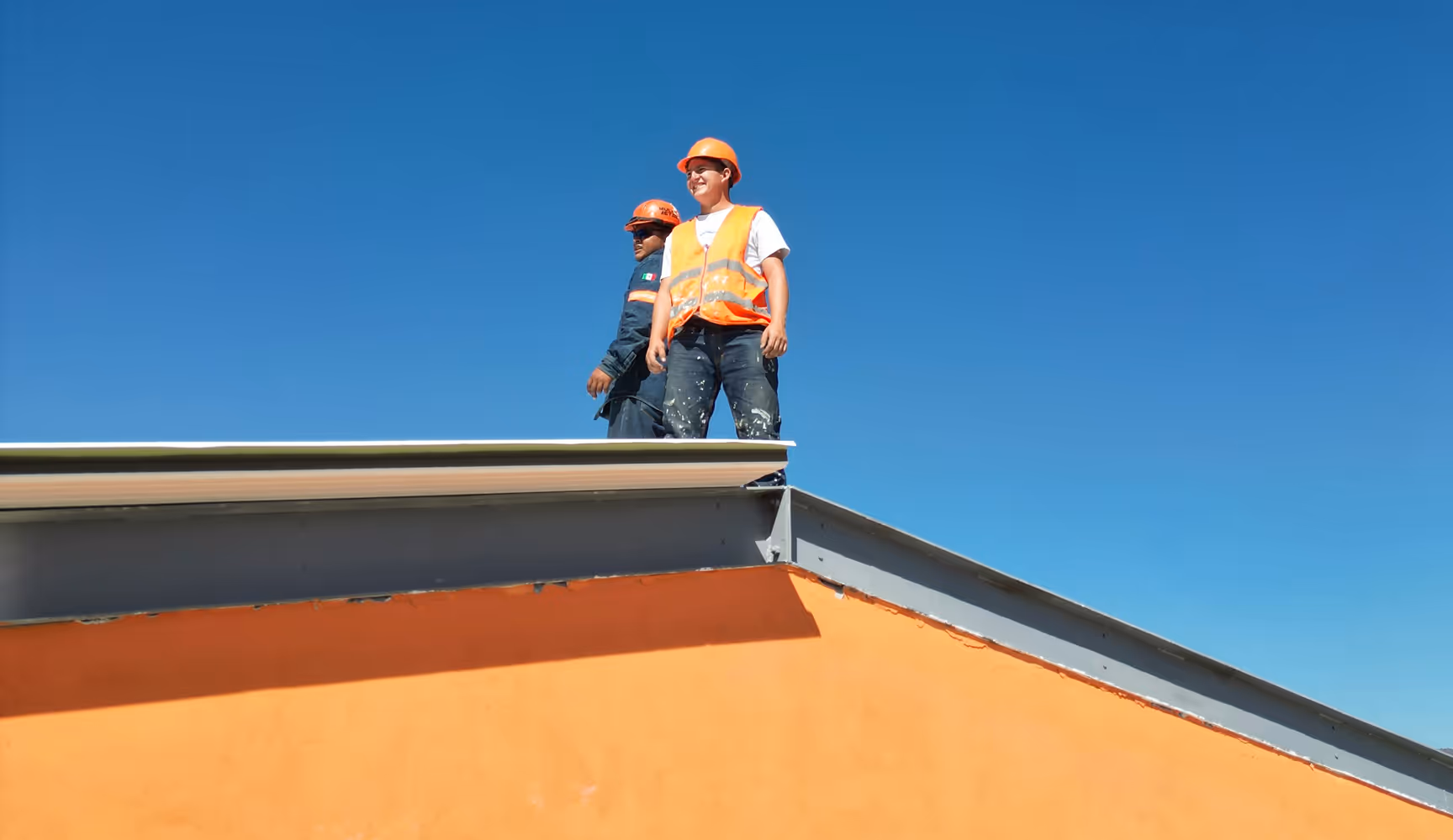 Two roofers stand at the edge of an orange-painted roof against a bright blue sky, representing the shingles, underlayment, and flashing Curri rushes to roofing contractors and supply yards on demand.