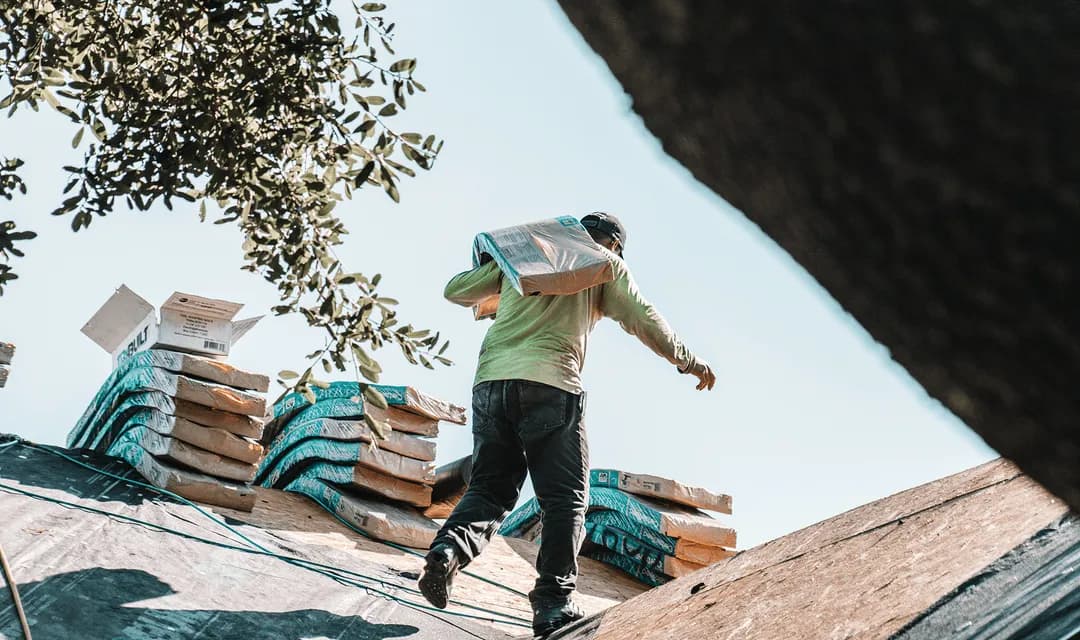 Roofer walks across a sloped roof with bundles of asphalt shingles stacked beside him, the kind of load Curri delivers same-day to roofing crews along with underlayment, flashing, and membrane.