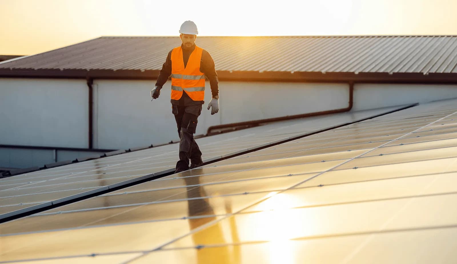 Solar technician in a safety vest walks a rooftop array of photovoltaic panels at sunset, representing the panels, inverters, and racking Curri delivers to solar installers and distributors nationwide.