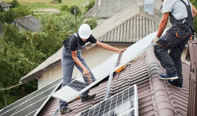 Two solar installers in hard hats and harnesses mount a photovoltaic panel onto a tile roof rail system, the kind of install Curri supports with same-day delivery of panels, inverters, and racking.