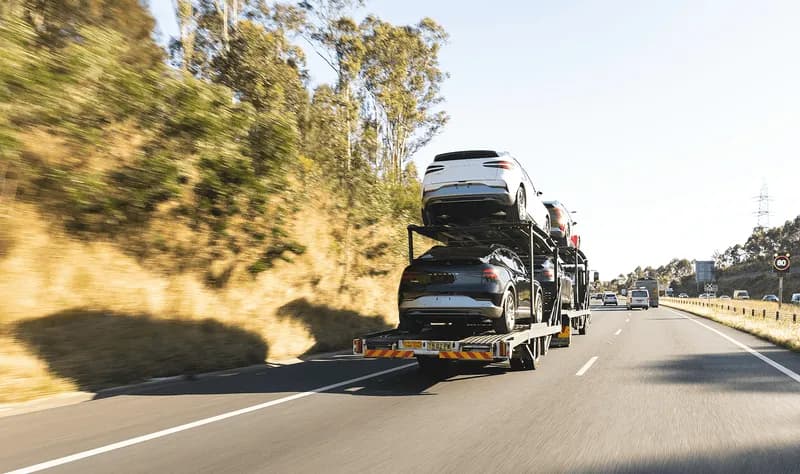Multi-car hauler truck loaded with vehicles cruises down a tree-lined highway, the kind of dealer-to-dealer, auction, and wholesaler transport Curri coordinates with local drivers nationwide.