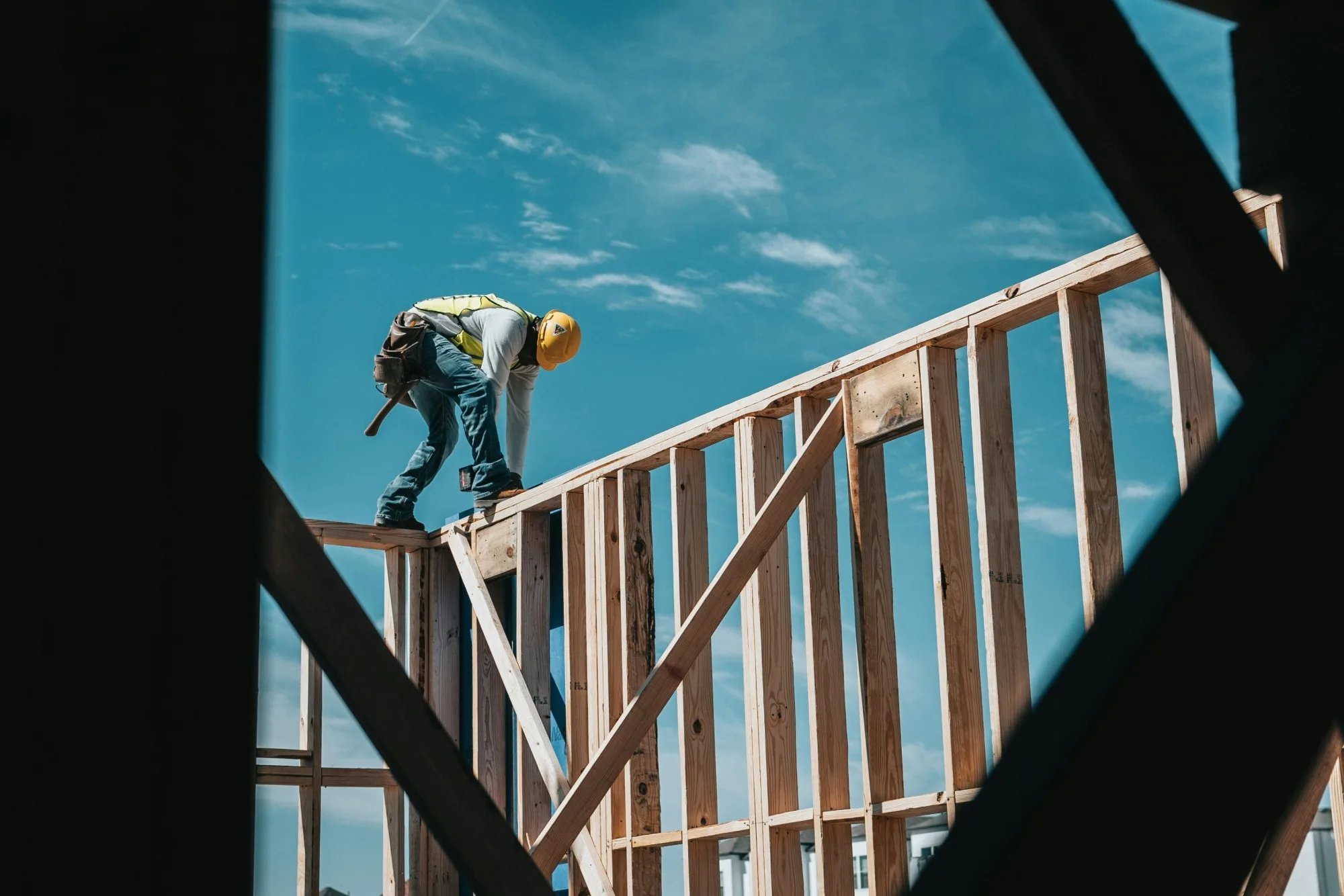 A framer in a hard hat working on top of a wood-framed wall against a blue sky