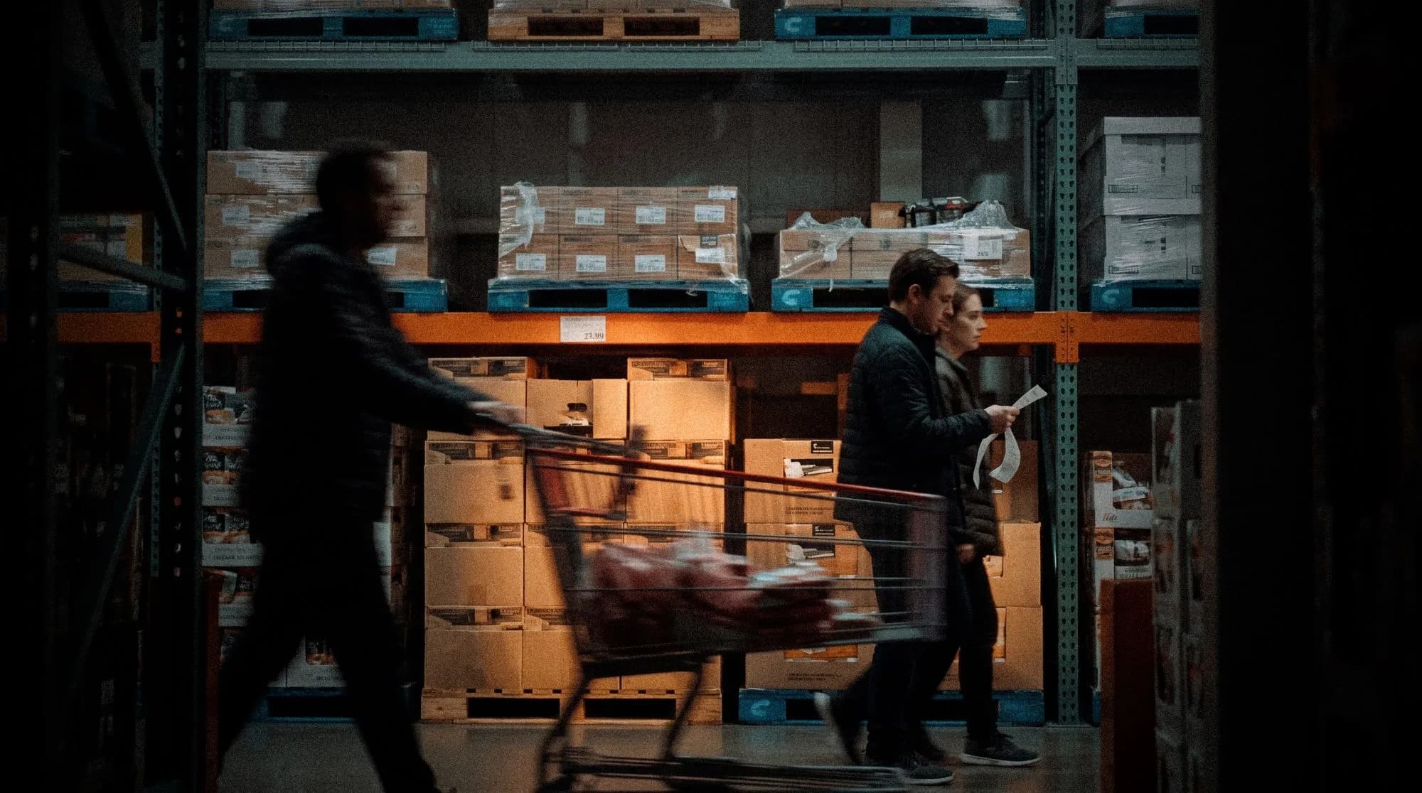 Shoppers pushing a cart past pallet racks of boxed goods in a warehouse club