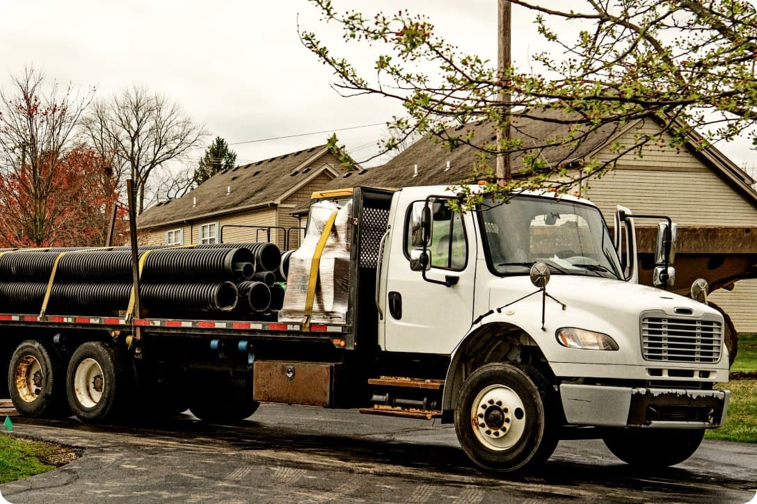A white flatbed truck loaded with strapped black corrugated drainage pipe parked on a residential street, with houses and budding trees behind it