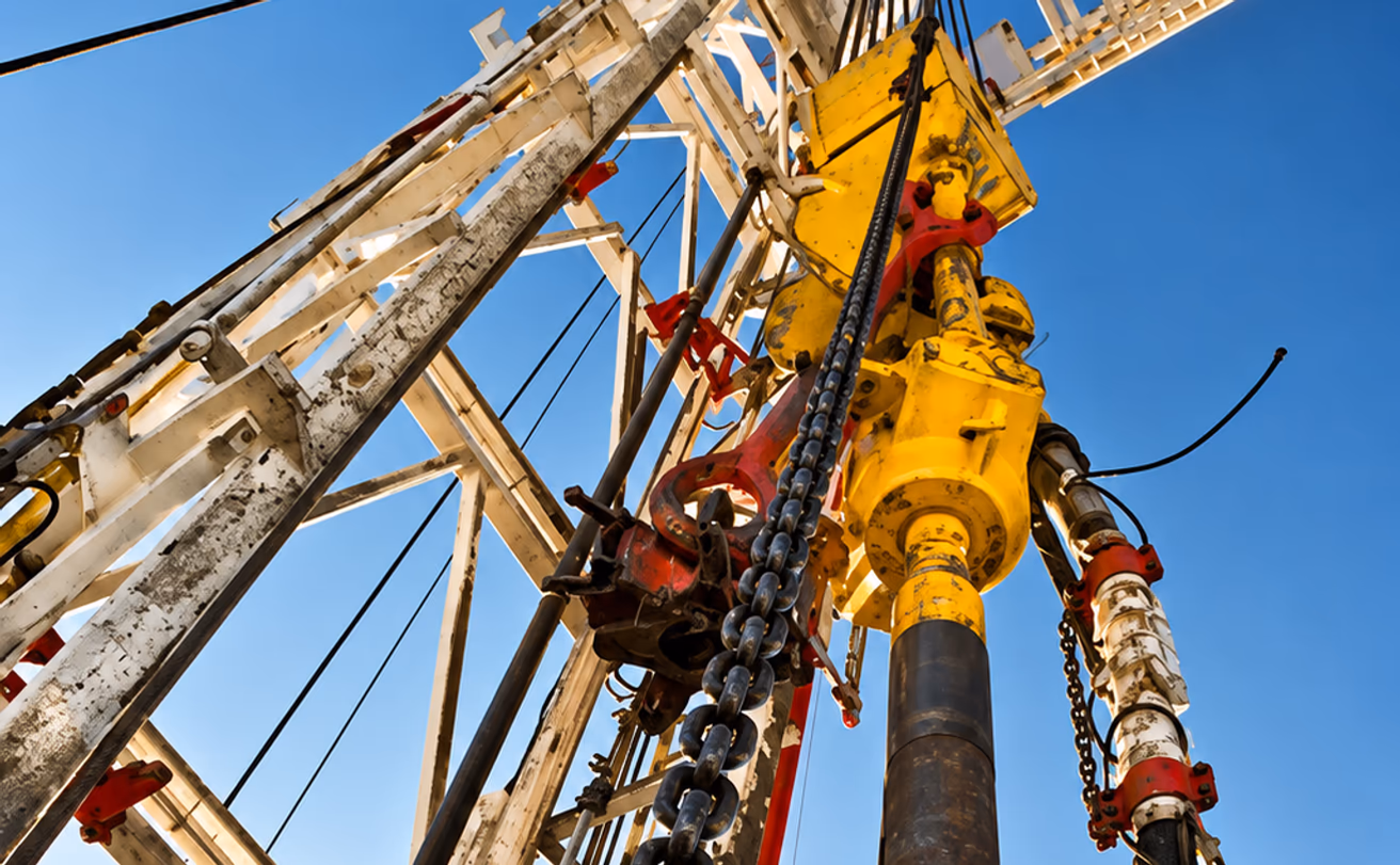 Looking up the mast of a drilling rig at the yellow top drive, tong chain, and drill pipe against a clear sky — the rig that goes down when a part is late and Curri runs hotshot to keep turning.