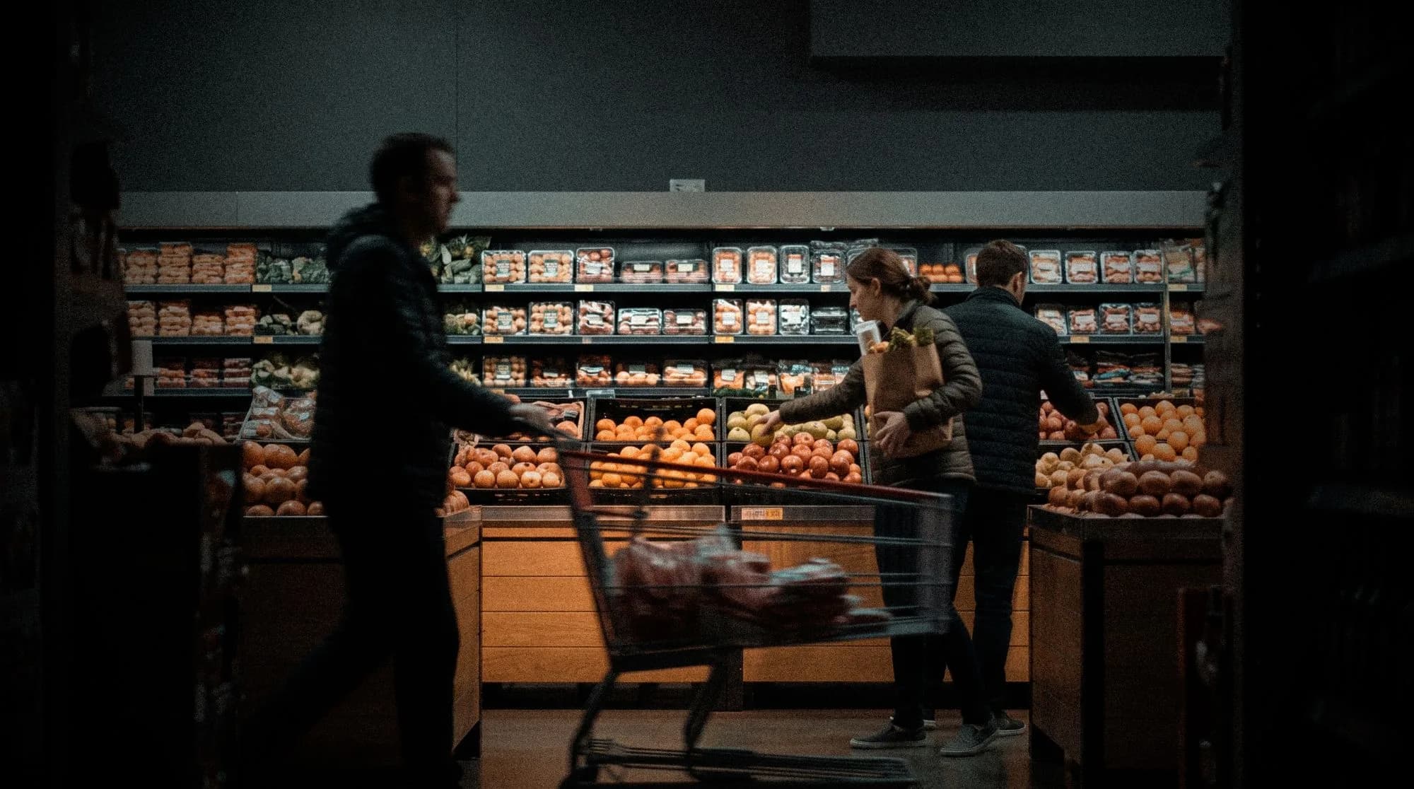 Shoppers in a grocery produce aisle, one pushing a cart past bins of fruit