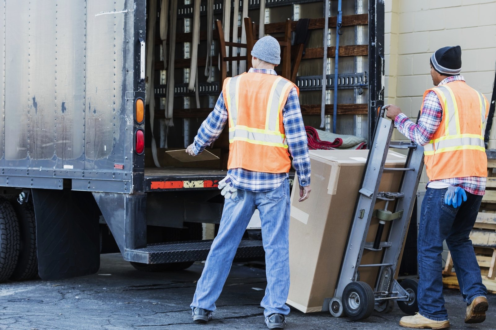 Two workers in orange safety vests unloading a large boxed item from the back of a box truck with a hand truck
